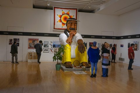 A large sculpture of a Native American man in white shirt and yellow pants sits in a room surrounded by spectators and other artworks