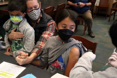 A 9-year-old girl with her sleeve rolled up and a bandage on her arm after getting vaccinated. She is holding her mom's hand and looking toward the syringe.