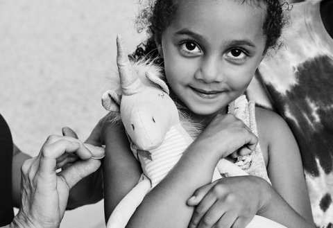 Black and white picture of a little girl holding a stuffed unicorn and getting her arm swabbed