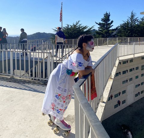 Latinx woman with hair in braids leaning on a bridge railing and looking out. She is wearing a white dress, white roller skates and Day of the Dead makeup.