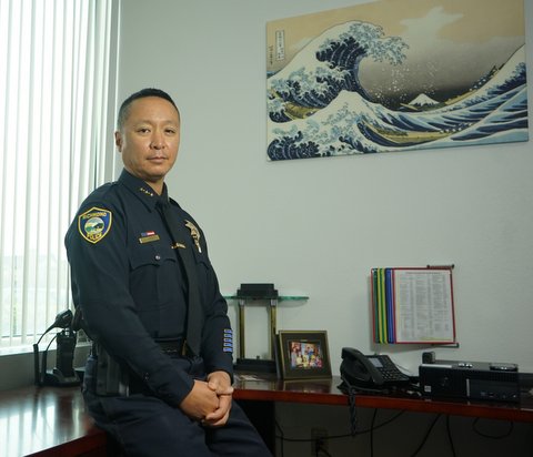 An Asian American man in a police uniform in an office with a painting of a wave on a wall.