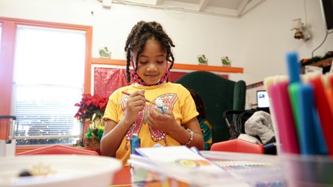 A smiling little Black girl painting a figurine. Her hair is in braids, and she is wearing a yellow T-shirt with a butterfly.