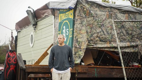 A middle-aged white man with a cigarette on his lip stands in front of a makeshift shelter made that features a green Oakland Athletics blanket