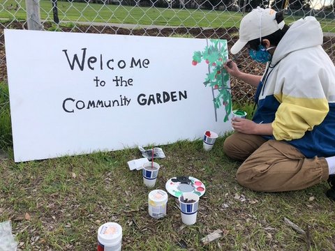 A person kneeling with art supplies and painting a plant on a sign that reads "Welcome to the Community Garden"