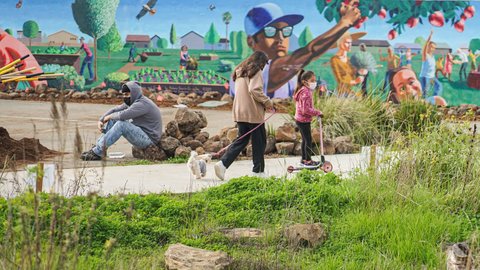 A woman walking a dog, a little girl on a scooter and a young man sitting on rocks in front of a mural.