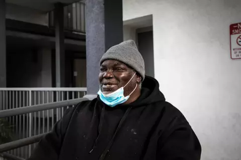 A smiling Black man wearing a beanie and a surgical mask pulled down to his chin