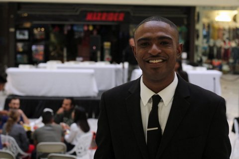 Black man in a suit. Other people can be seen sitting at a table in the background
