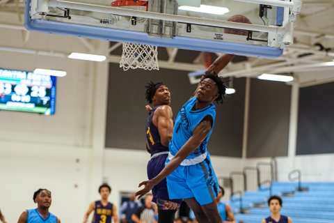 A basketball player moves to dunk the ball with an opponent jumping alongside him