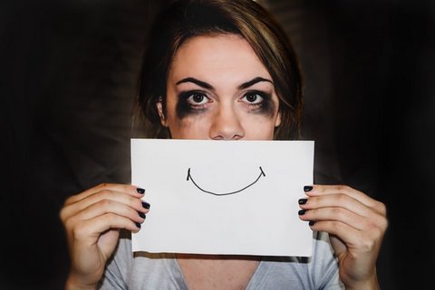 Woman with dark makeup smudged around her eyes holding up a drawing of a smile in front of her face