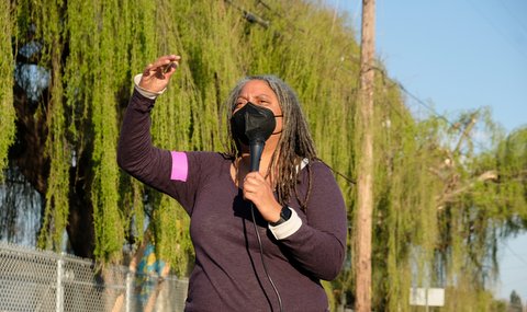 Black woman wearing mask and holding microphone looking off into the distance with her arm raised, facing palm down, and trees behind her