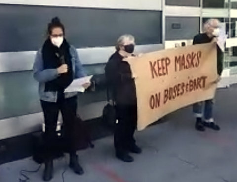 Masked person with microphone next to two older people holding a banner that says "keep masks on buses and BART"