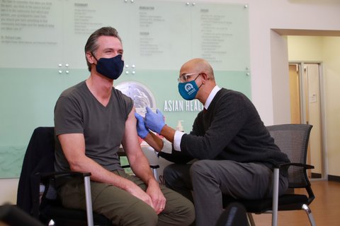One man administering a vaccine shot to another. Both are wearing masks.