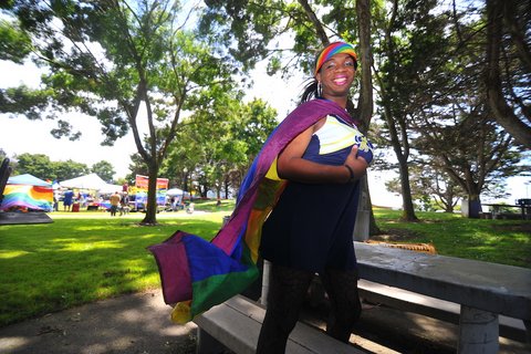 Black person wearing pink lipstick, rainbow headscarf and rainbow flag like a cape.