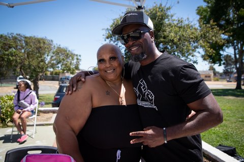A Black woman with shaved head and hoop earrings standing next to a Black man with his arm around her shoulders