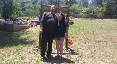 A man and woman standing in a field of mostly dead grass. He is making bunny ears behind her head and she is looking up at him. They are standing in front of a sign for a cemetary near a colorfully decorated area.
