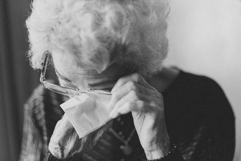 Older woman with head bowed, dabbing eye with tissue under glasses. In black and white