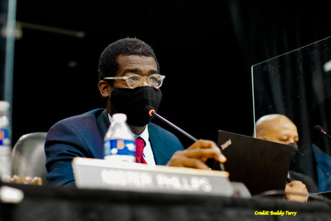 A Black man wearing suit, mask and glasses seated at a microphone