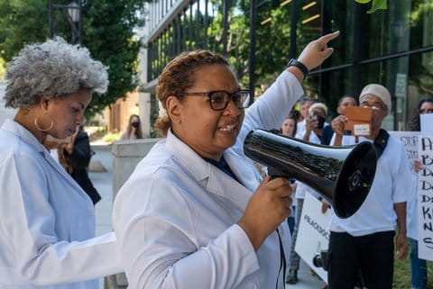 A Black woman wearing white lab coat and carrying a megaphone as part of a rally
