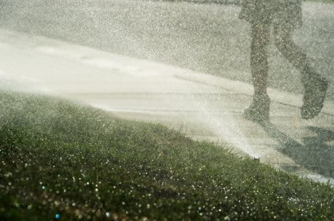 Ground-level view of sprinklers spraying water onto grass and in the air as person walks by