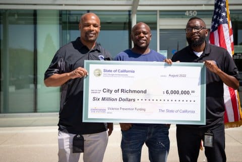 Three Black men holding an oversized check for $6 million from the state of California to the city of Richmond. The memo says "violence prevention funding"