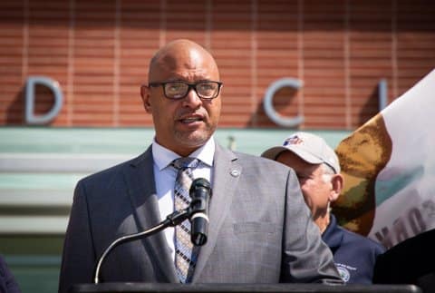 A bald Black man wearing a suit and glasses standing in front of a microphone