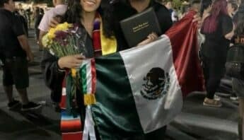 A young Latina woman dressed in cap and gown with her father. They are holding up a Mexican flag. She is also holding a bouquet of flowers and her diploma holder.