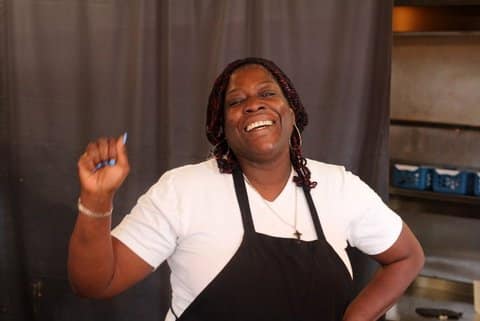A Black woman with a joyous look on her face and one fist raised. She is wearing blue nail polish and a black apron.