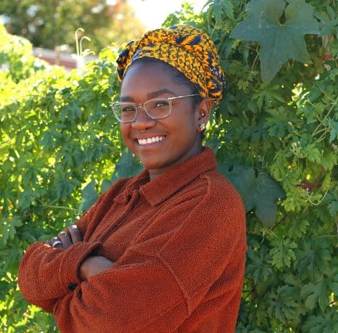 A smiling young Black woman wearing glasses and a headscarf standing with her arms folded in front of tall greenery