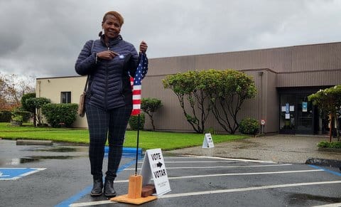 A Black woman with short hair and a puffer jacket stands next to a small American flag and a vote sign outside of her polling place. She is pointing to a sticker on her coat. It is likely an I voted sticker, but it is not legible in the photo