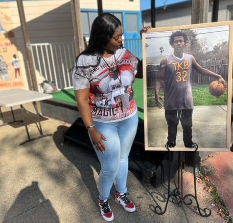 A young Black woman standing next to and looking at a photo of her brother