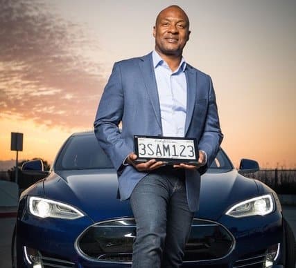 A Black man holding a digital license plate in front of an expensive-looking car. The plate is on a tablet The display looks like a regular license plate.
