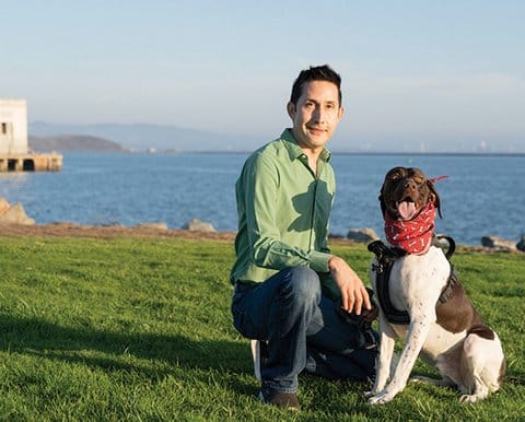 A Latino man kneeling next to a brown and white dog wearing a red bandanna on grass in front of the San Francisco Bay.