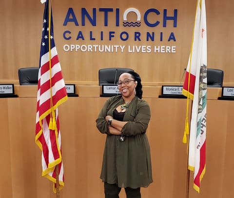 A Black woman standing between U.S. and California flags in government chambers in front of wall that says Antioch California Opportunity Lives Here