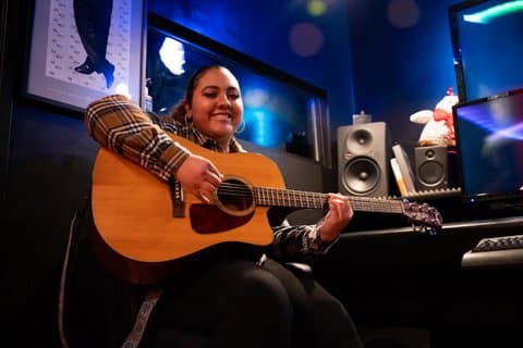 A smiling young Latina woman seated with an acoustic guitar looking down toward the camera