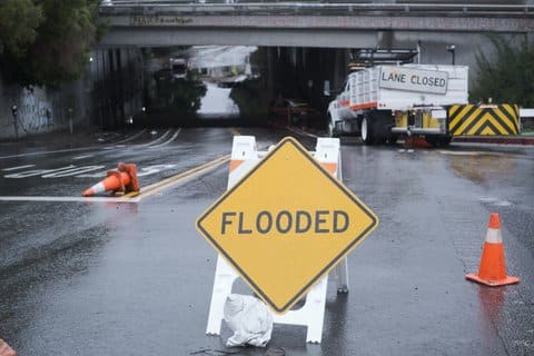 A sign that says "flooded" in front of a closed off street. Also on the street are a few orange cones and large truck with a sign that says "lane closed"