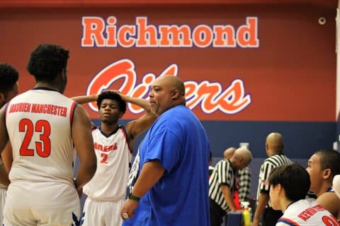 A heavyset Black man, who is a high school basketball coach, with some of his players during on the sidelines during a game. On the wall in the background, it says Richmond Oilers