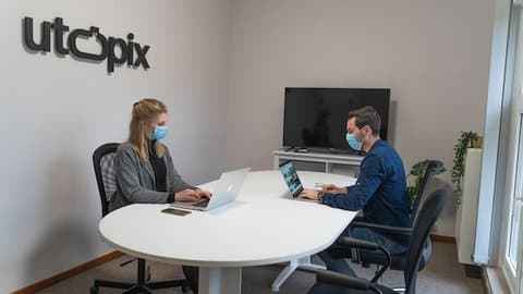 A woman and man at a table, both wearing surgical masks and working on laptops. A large monitor is facing them, turned off. A sign on the wall says utopix.