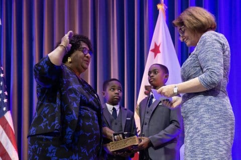 Shirley Weber, a Black woman with one hand on a book and one raised as she sworn into office by Toni Atkins, a white woman. Standing by are Weber's grandsons, two young Black boys in suits. One of them is holding the book.