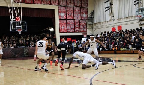 Scene from a high school boys basketball game with one player on the ground after diving toward a loose ball and opponents and teammates looking on.