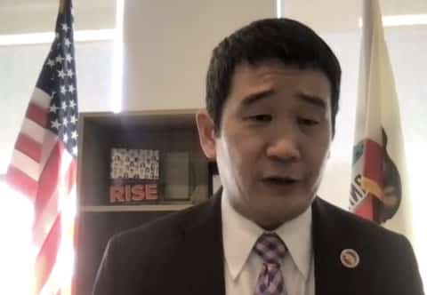An Asian man in a suit looking down. U.S. and California flags are behind him.