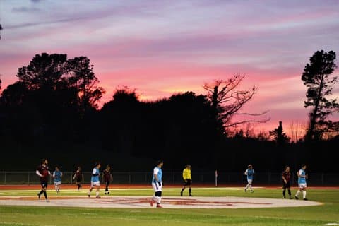 Wide shot of a soccer game at sunset. The sky is awash in purplish and pinkish tones.