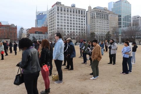 Several people standing in a sandy open area with tall buildings in the background. Some people's heads are bowed.