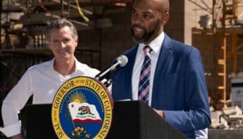 Antioch Mayor Lamar Thorpe, a Black man, stands at a lectern with the Seal of the Governor of the State of California. Looking on with a smile is Gov. Gavin Newsom.