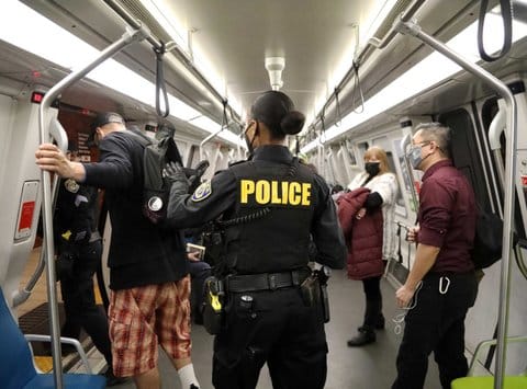 Inside a BART train car. People are wearing masks. In the center of the photo is a police officer seen from behind. The officer appears to be a Black woman.