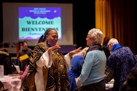 A Black woman and white woman engaging with each other at an event. In the background, a screen says "Welcome Bienvenidos"