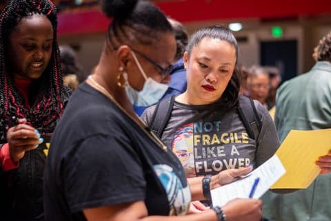 Two women at an event in an auditorium looking at sheets of paper. One is wearing a surgical mask. The other is wearing a T-shirt with the words "not fragile like a flower." Another woman is near them.