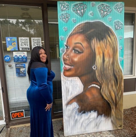 A Black woman in a blue velvet dress stands next to a large painting of a young Black woman