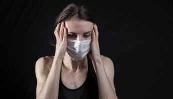`A white woman wearing a medical face mask has a distressed expression and is holding her hands to the sides of her head