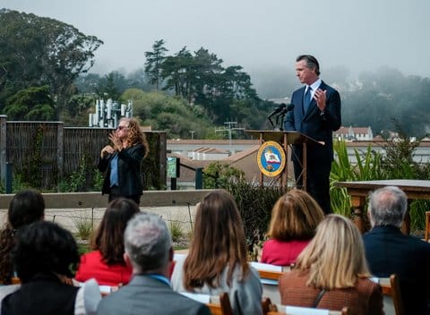 View looking over seated audience from behind toward Gov. Gavin Newsom standing at a lectern with trees in a hilly and foggy area behind him