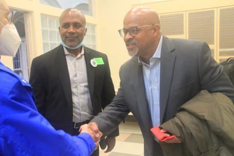 Two Black men in suits and an older person in a mask. One of the men is shaking the older person's hand